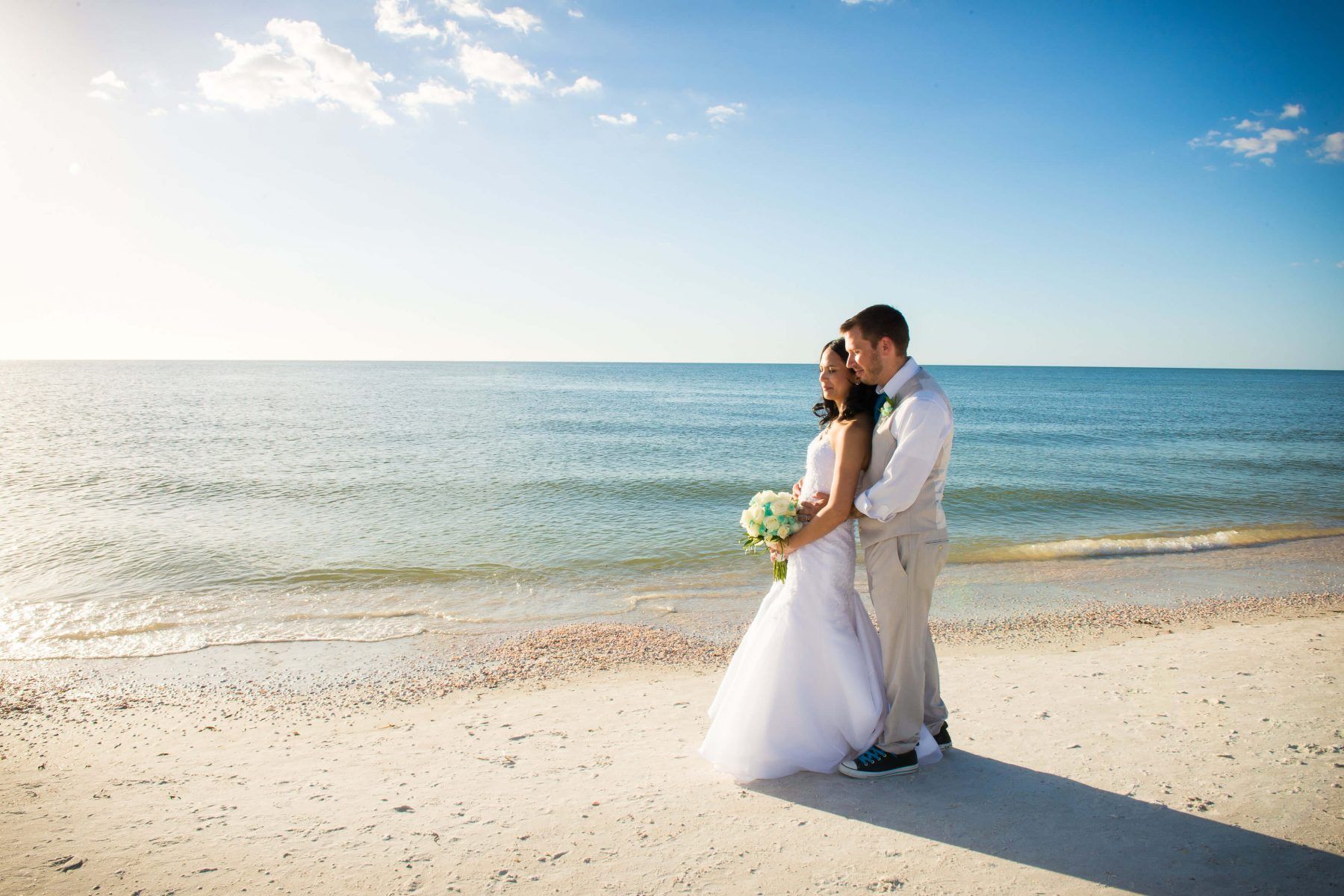 Bride and groom embrace on a sunny beach, holding a bouquet. Calm blue ocean and sky in background.