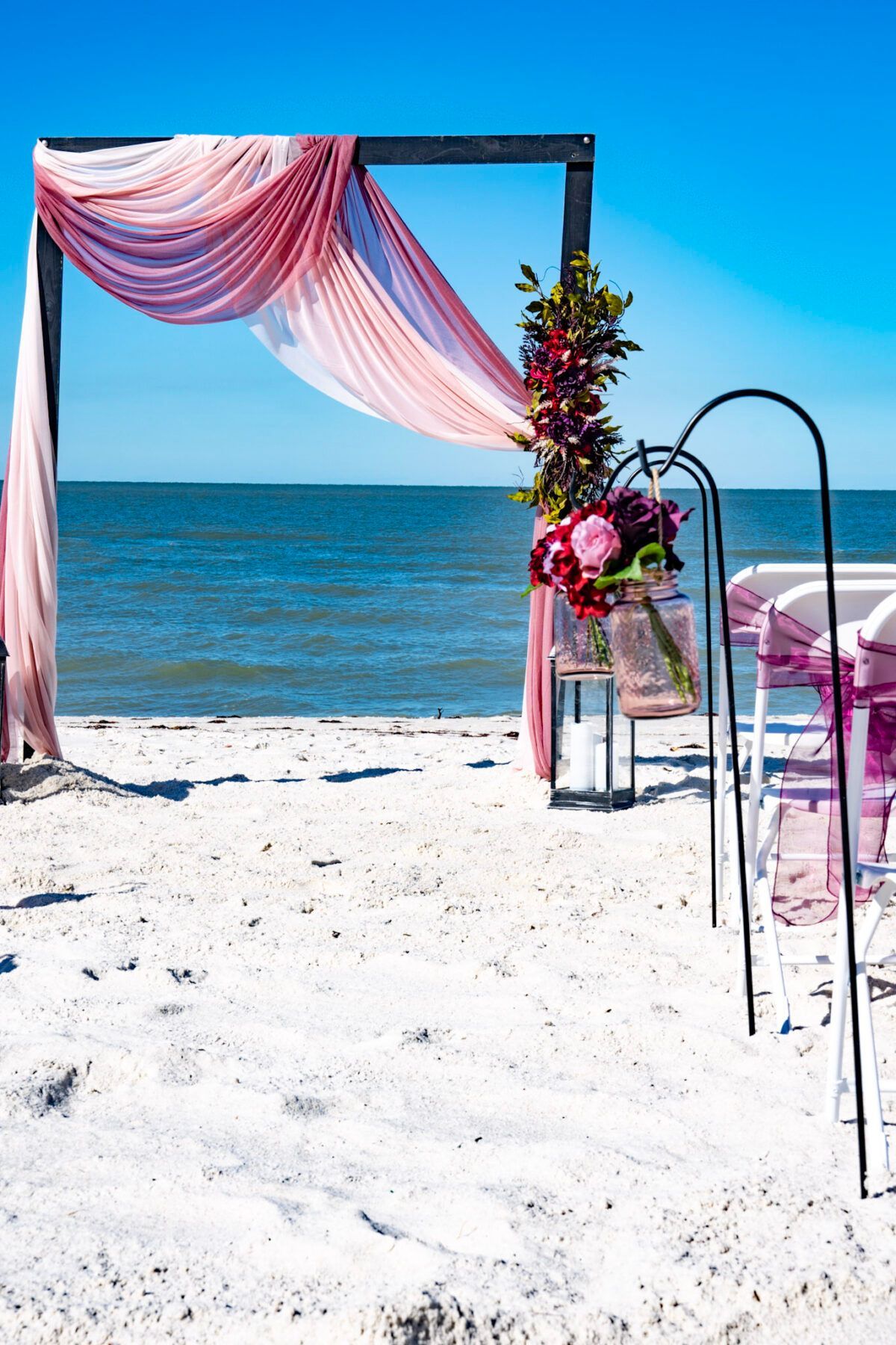 Beach wedding setup with draped pink fabric, flowers, and chairs against the ocean and blue sky.