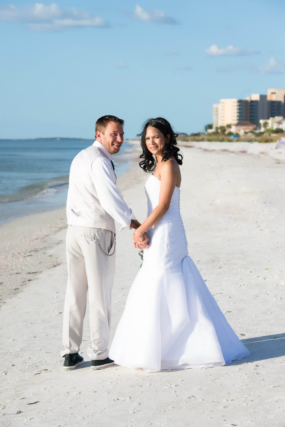 Bride and groom holding hands on a white sandy beach, smiling, wearing wedding attire.