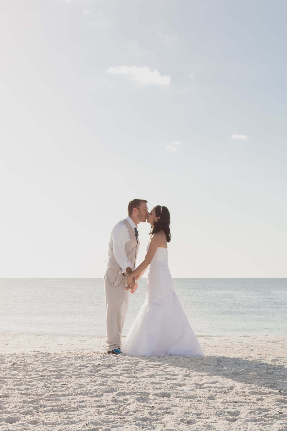Couple kissing on a beach; the bride wears a white dress, the groom tan pants and vest, ocean in background.