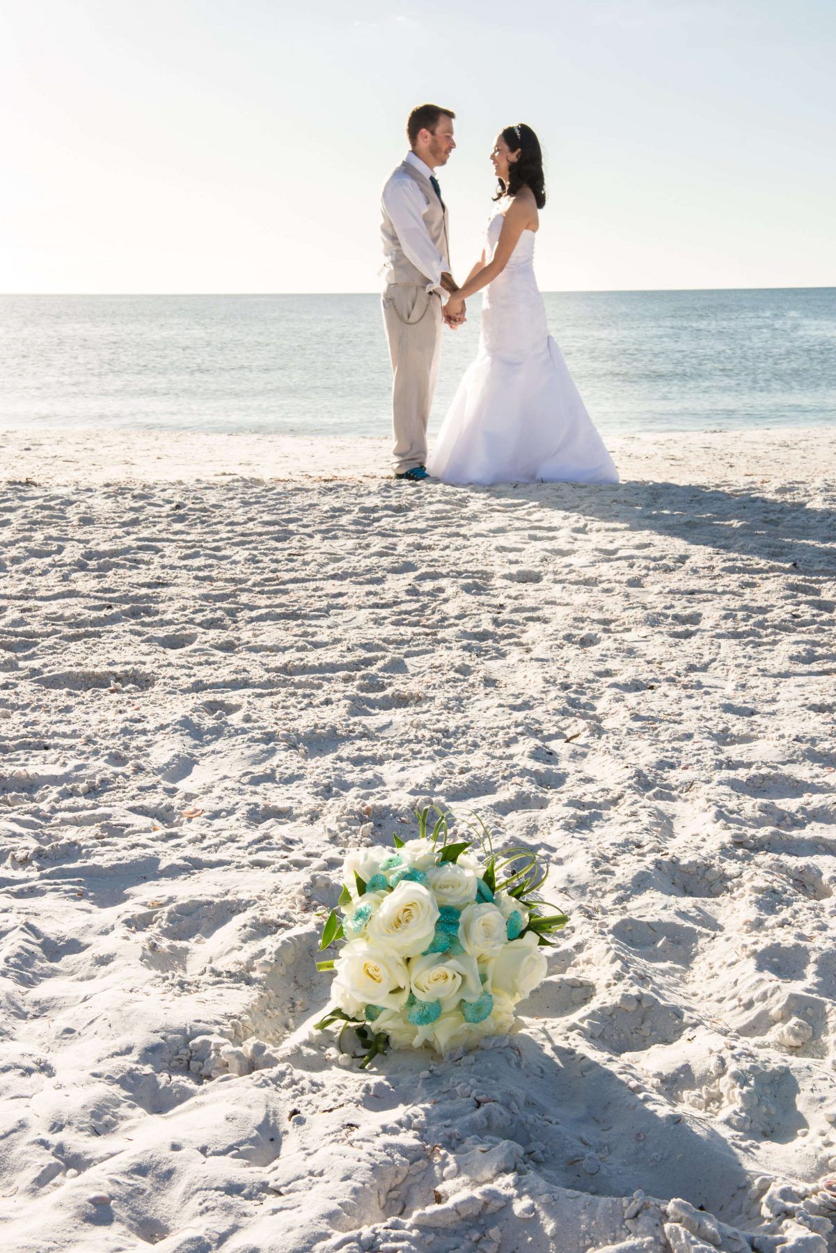 Wedding couple on a beach holding hands, bouquet in foreground. Sunny day, white sand, clear water.