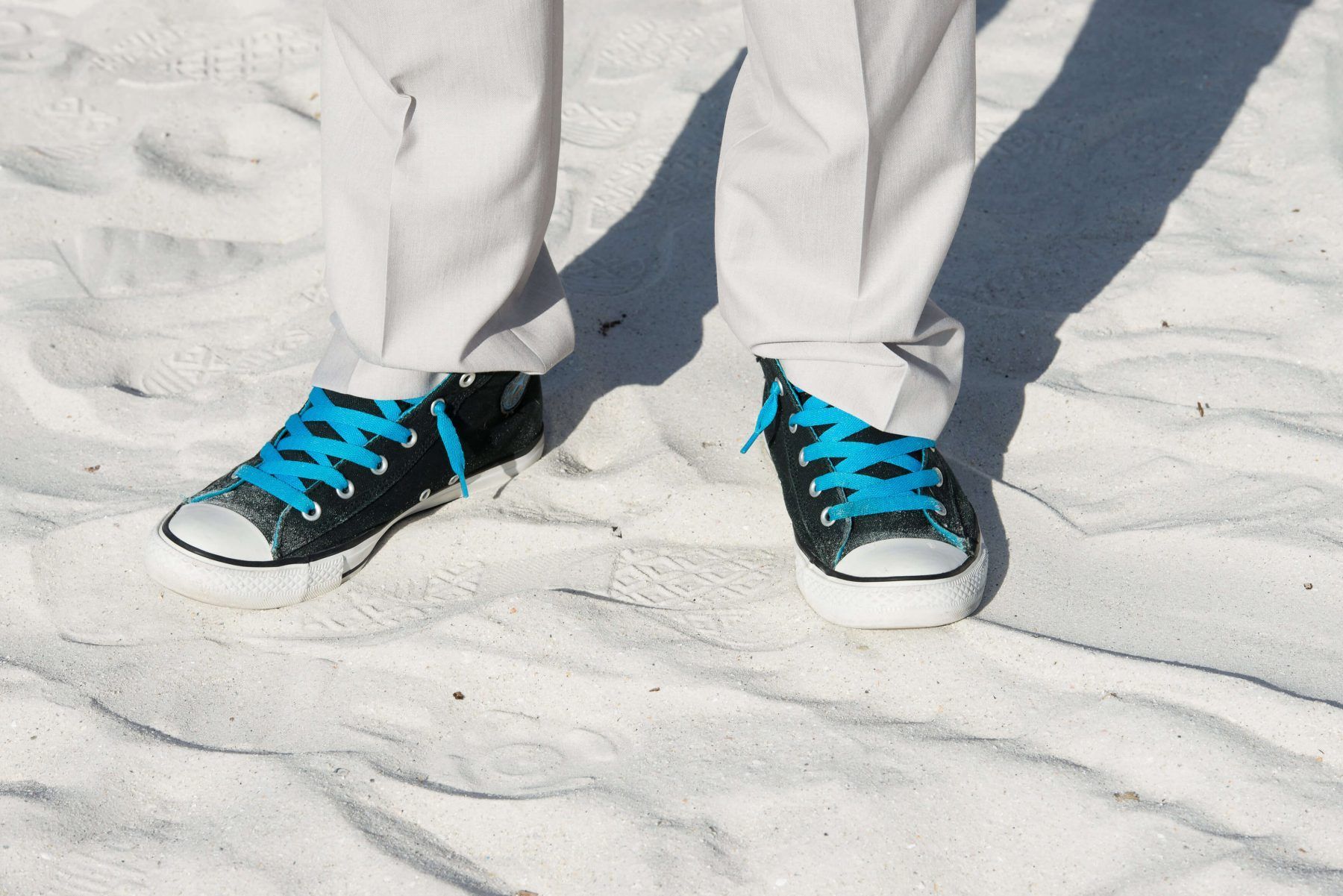 Person's legs in white pants and black sneakers with blue laces, standing in white sand.