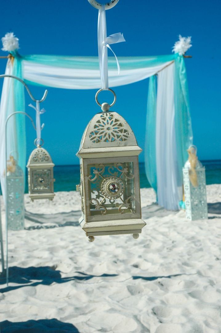 Beach wedding decor: Lanterns, draped fabric in shades of white and teal. Sunny day, bright blue sky.