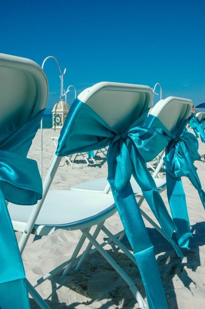White folding chairs with turquoise sashes arranged on a sandy beach under a clear blue sky.