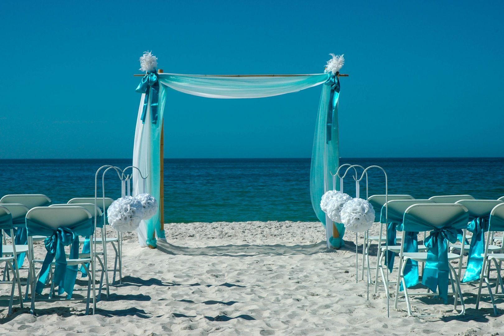 Beach wedding ceremony setup: arch with turquoise fabric, white chairs, ocean backdrop.