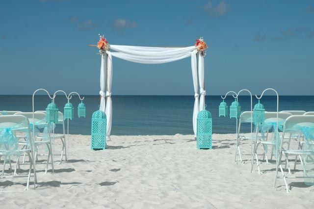 Beach wedding ceremony setup: white chairs, turquoise accents, draped arch, ocean backdrop.