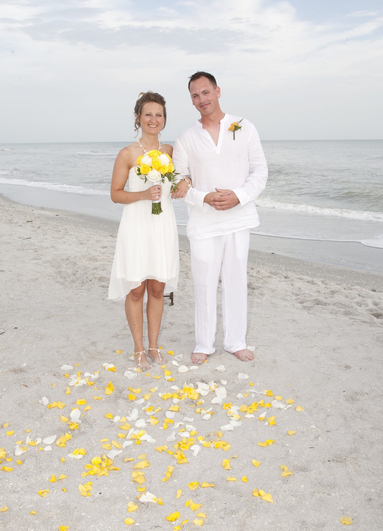 Couple in white wedding attire on beach, holding hands, smiling. Yellow petals on sand.