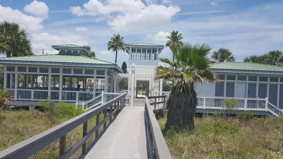 Wooden boardwalk leading to a white, open-air building with a central tower on a sunny beach.