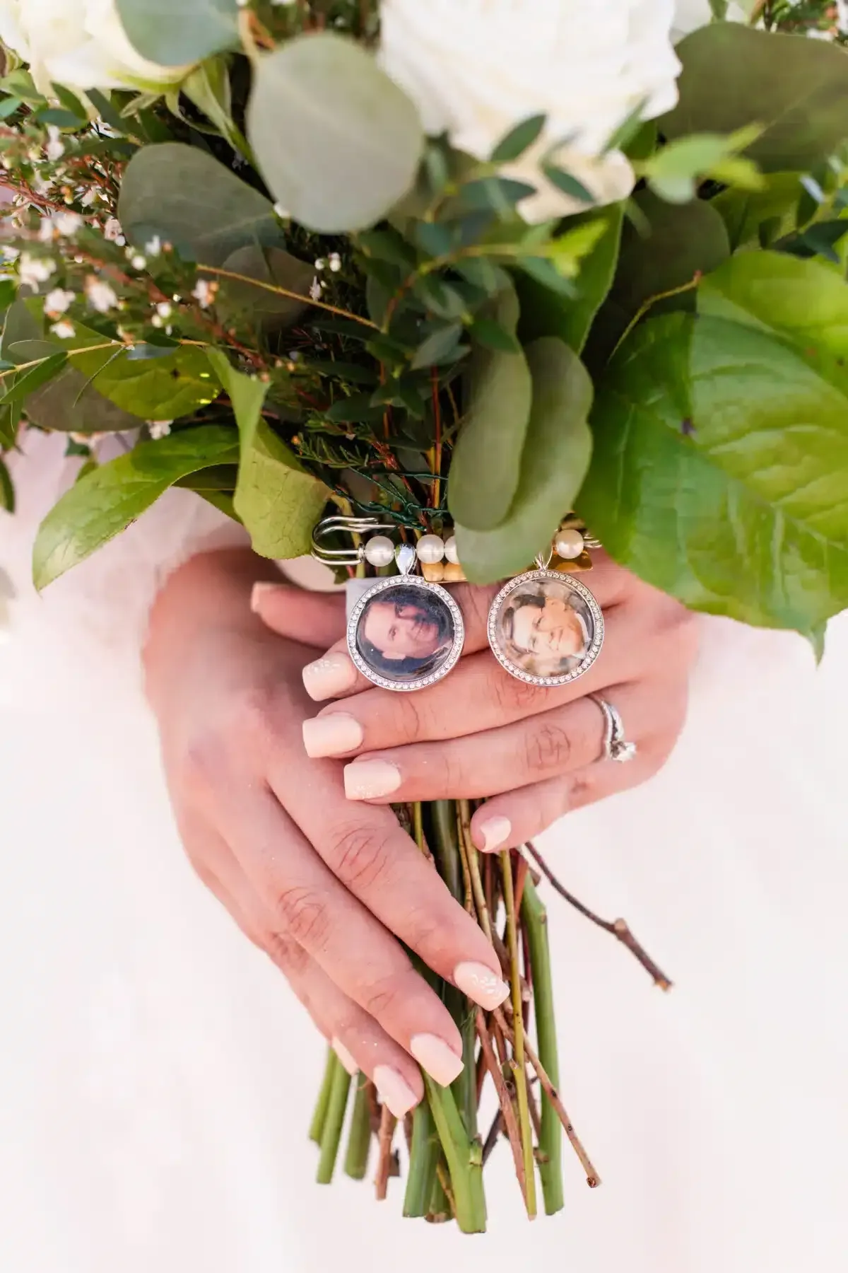 Bride holding a bouquet with photo charms. Beige nails, white dress, green foliage, and white flowers.