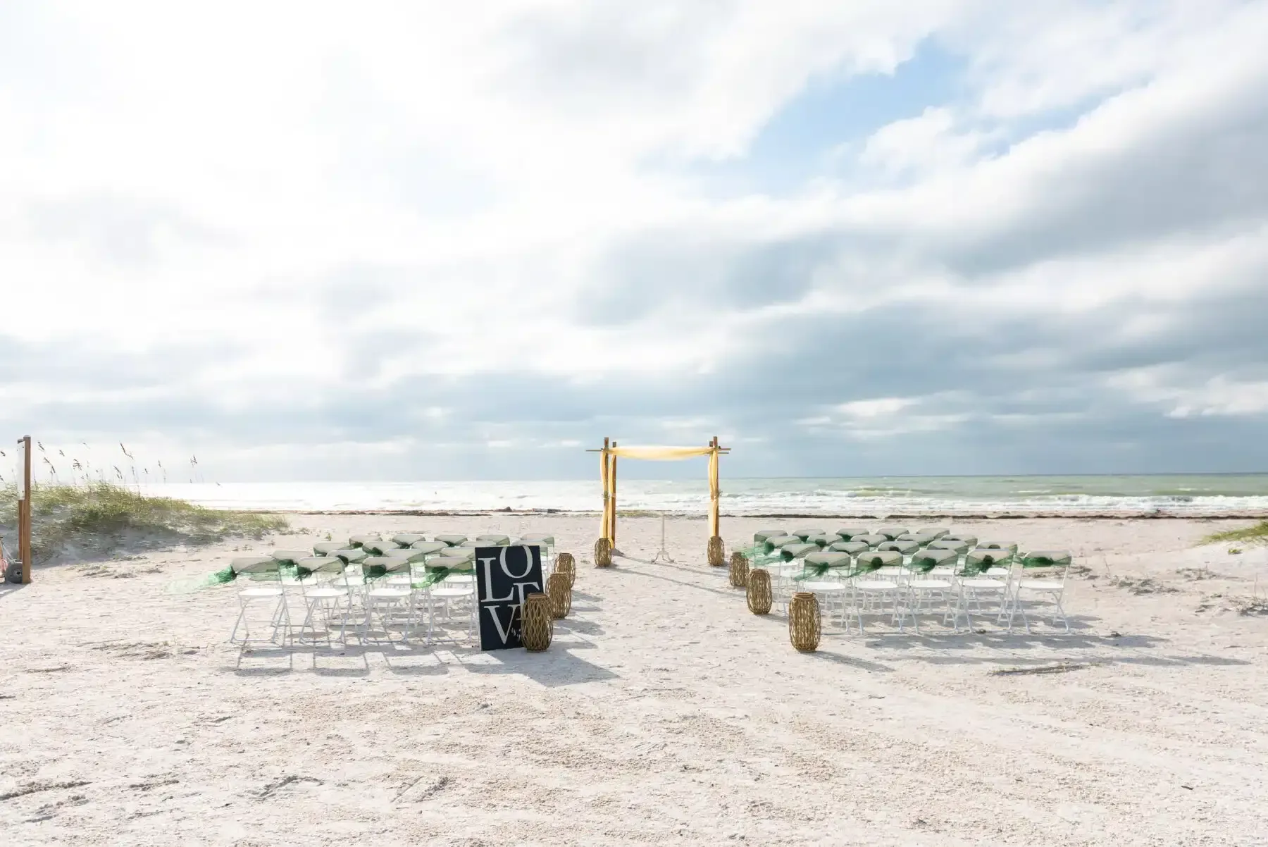 Beach wedding ceremony setup: rows of clear chairs face a decorated arch on a sandy shore under a cloudy sky.