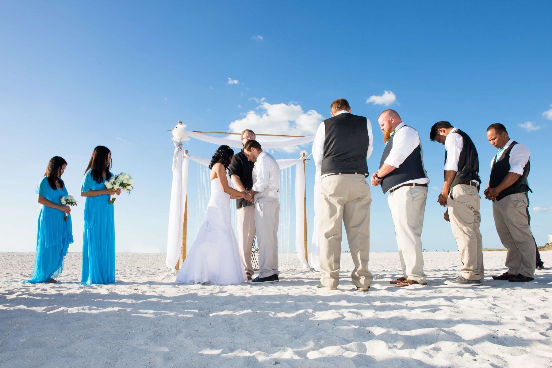 Wedding ceremony on a white sandy beach with couple under a canopy, bridesmaids in turquoise, groomsmen in vests.