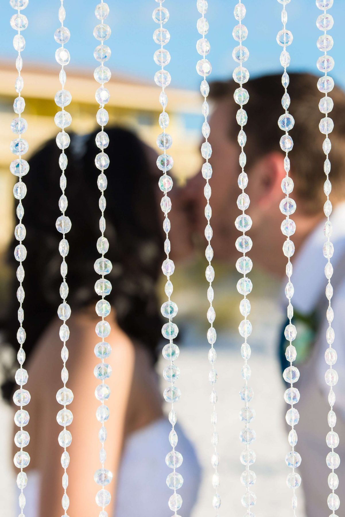 Wedding ceremony: Bride and groom kissing behind beaded curtain on beach.