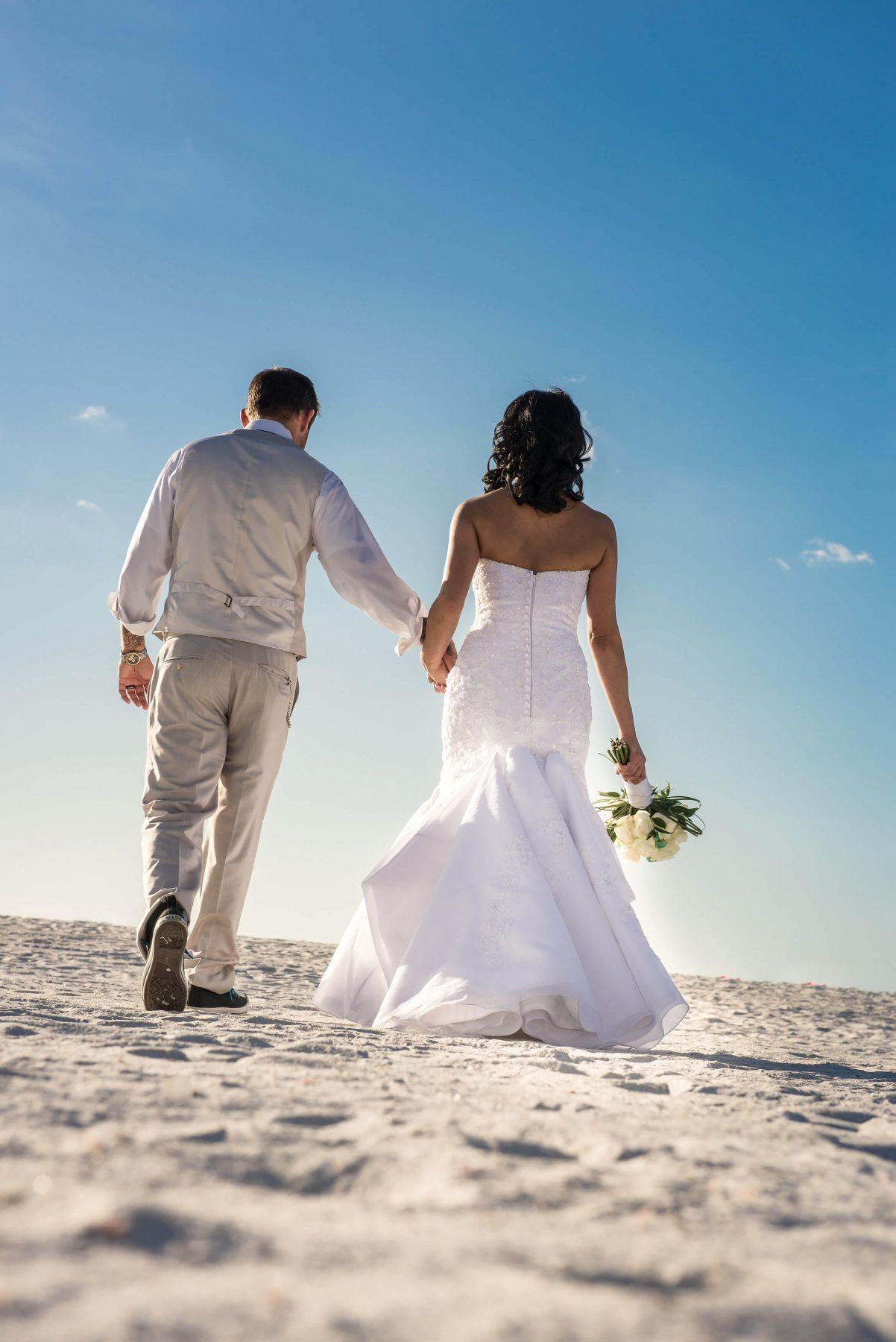 Bride and groom holding hands, walking on white sand towards bright blue sky.