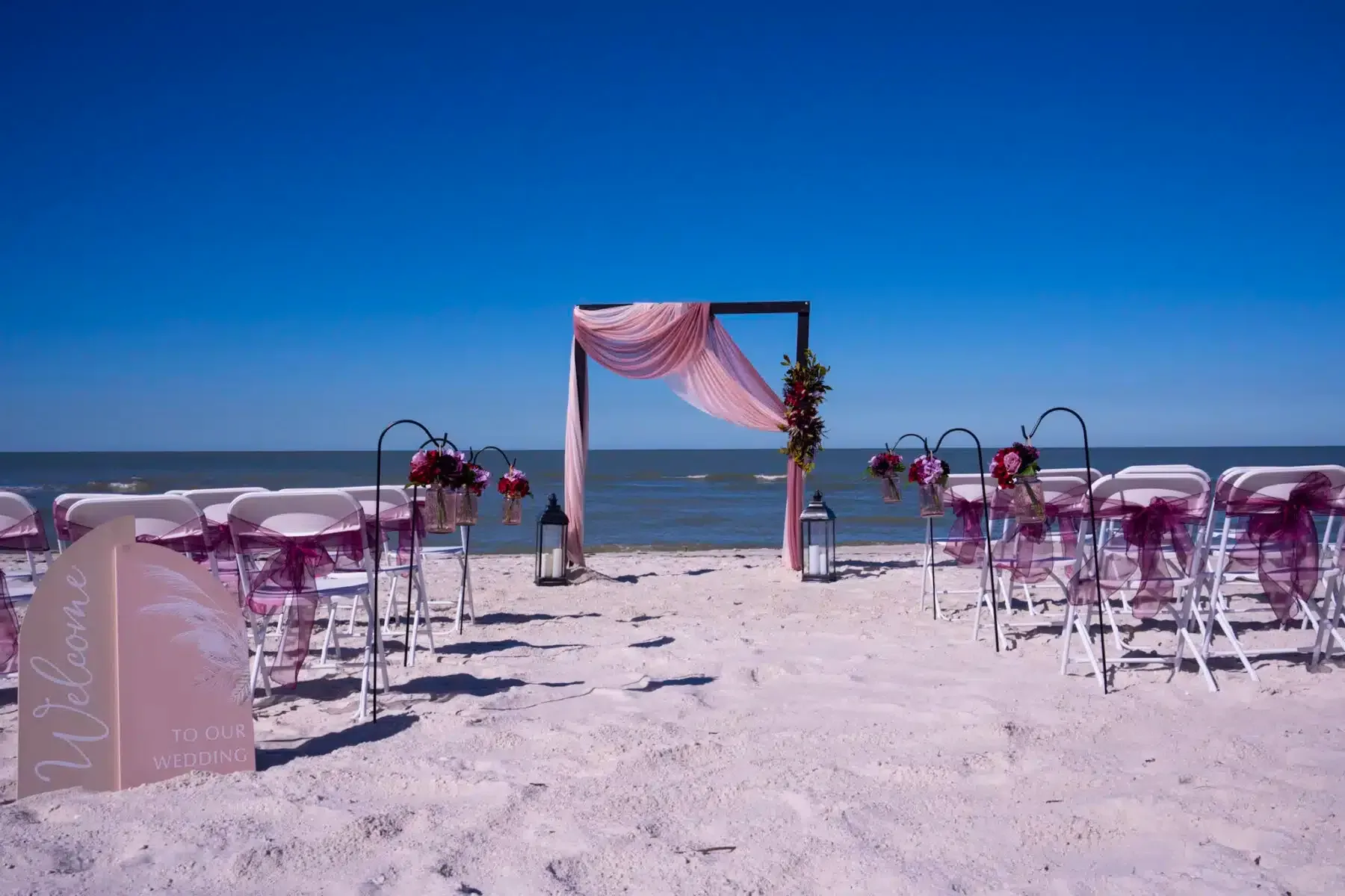 Beach wedding setup with arch, chairs, and decorations. Ocean and blue sky background.