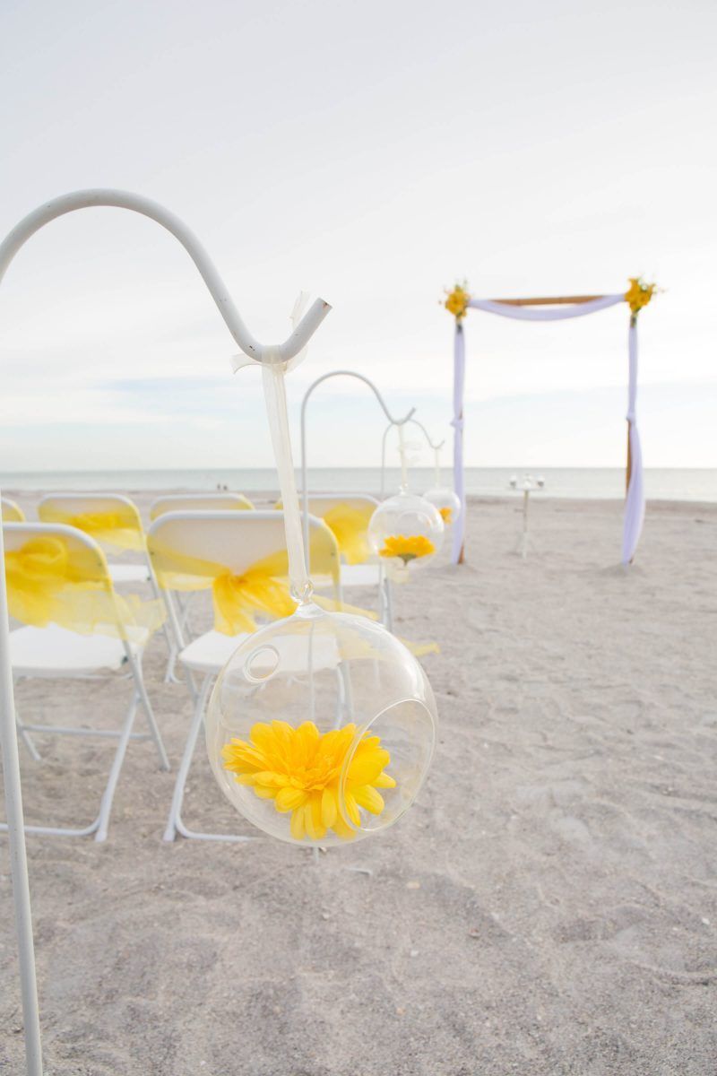 Beach wedding setup with yellow accents: chairs, flowers in glass orbs, and arch.