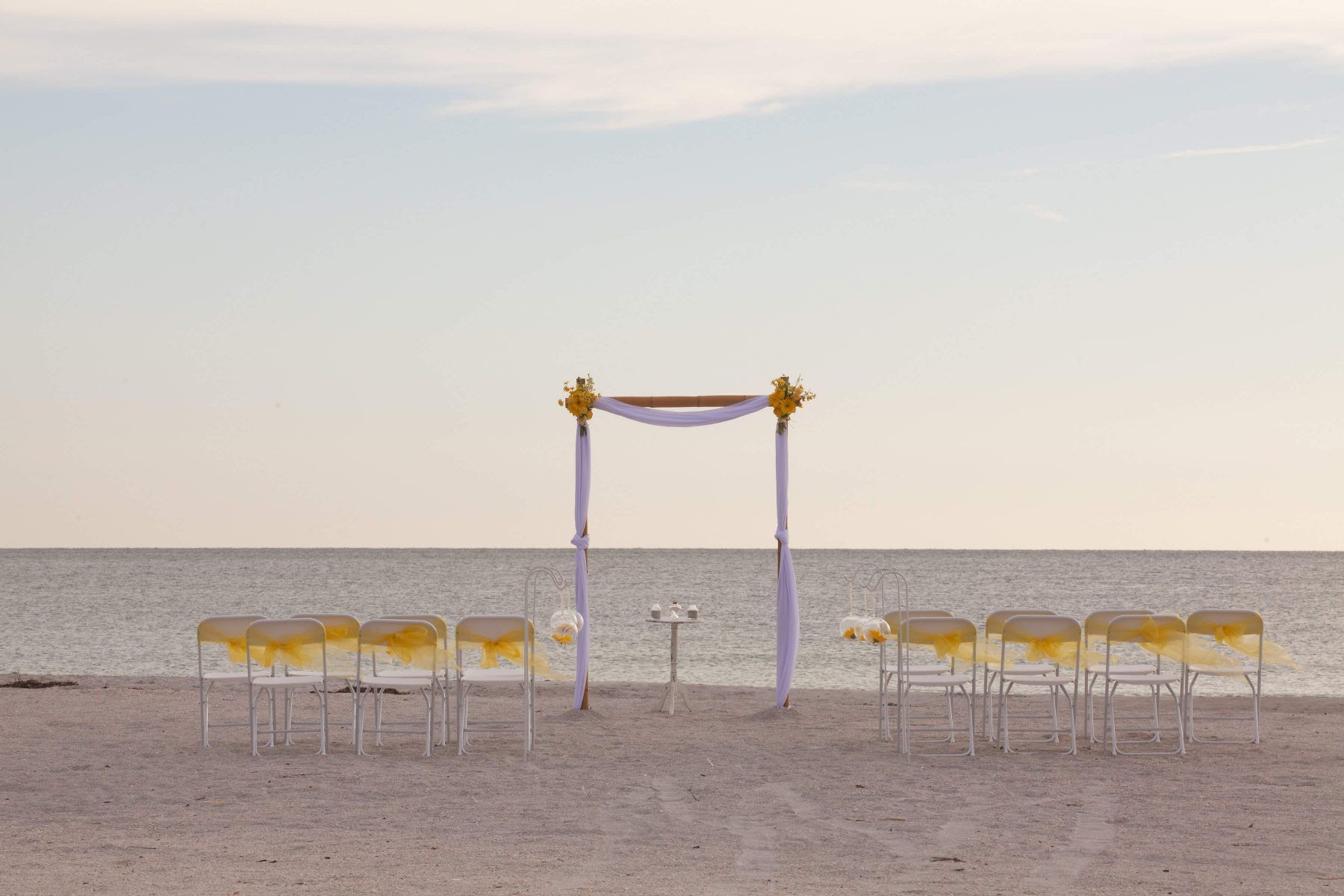 Beach wedding setup with chairs facing a decorated arch, ocean in background.