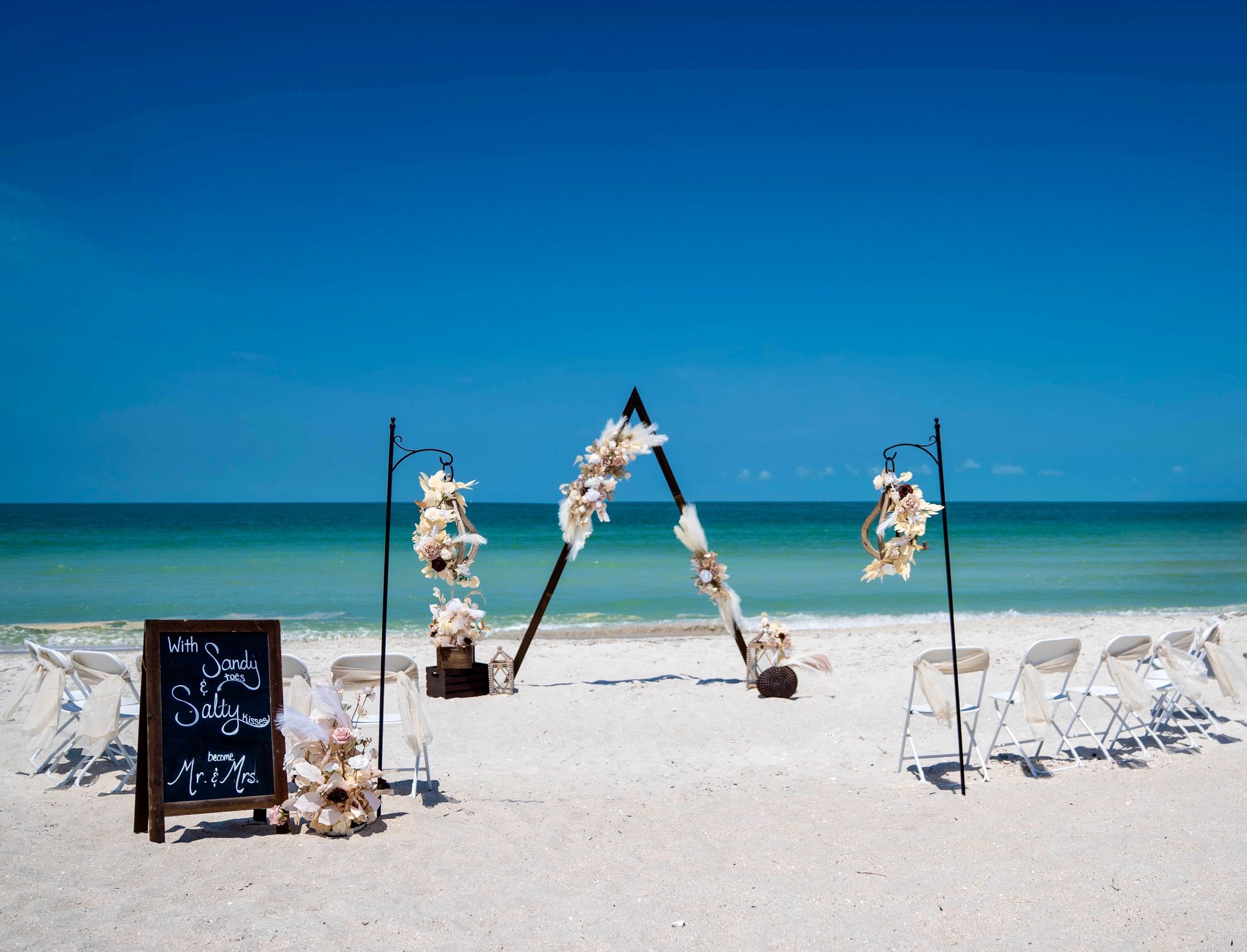 Beach wedding setup with an arch decorated with flowers and empty chairs facing the ocean under a clear blue sky.