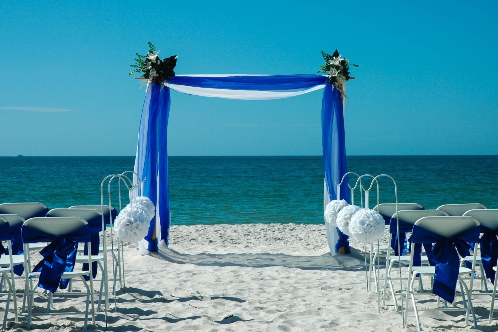 Beach wedding setup with blue draping, white chairs, and ocean backdrop.