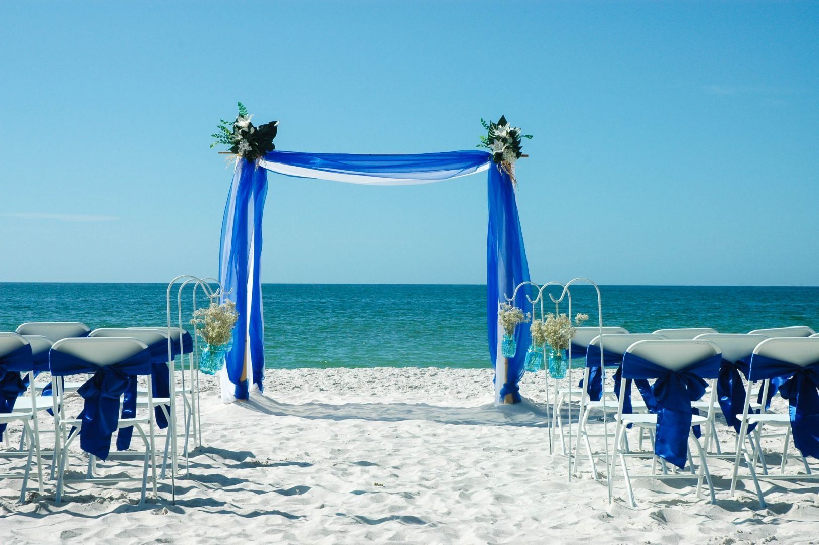Beach wedding setup: blue draped arch, white chairs, ocean backdrop.