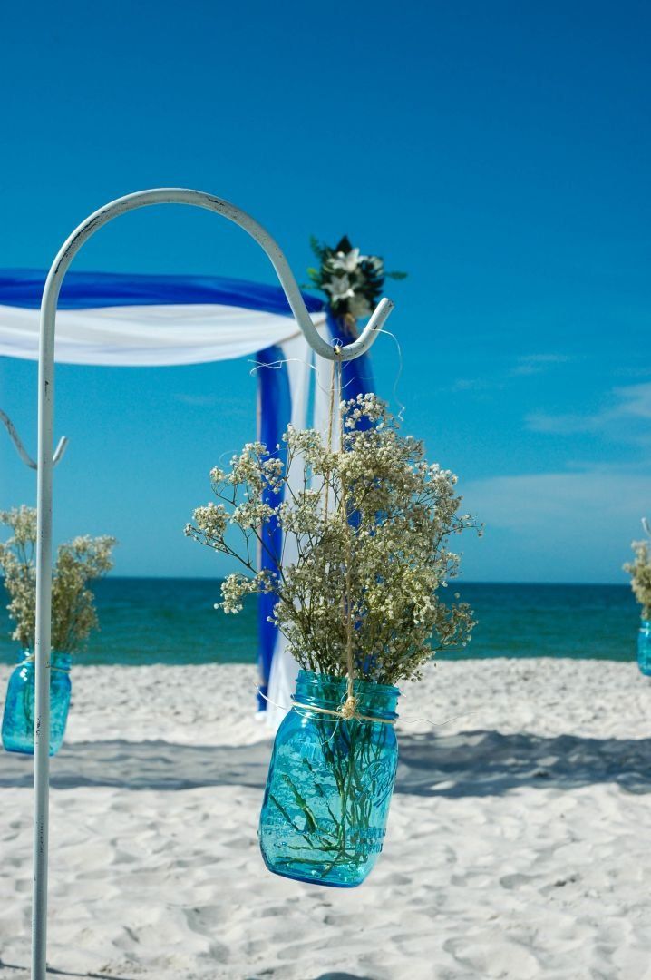 Blue mason jars with white flowers hang on a metal arch on a beach with blue and white fabric in the background.