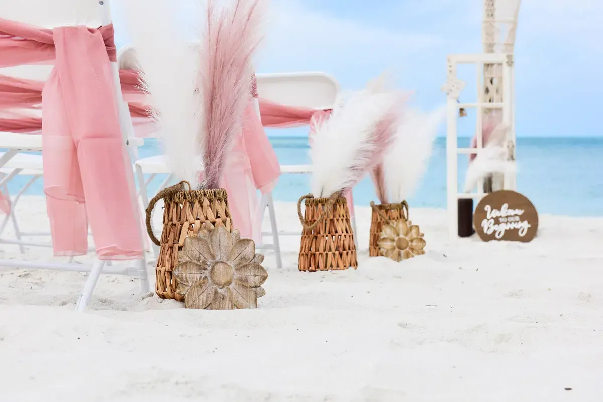 Beach wedding setup with chairs, pink draping, floral arrangements, and a sign.