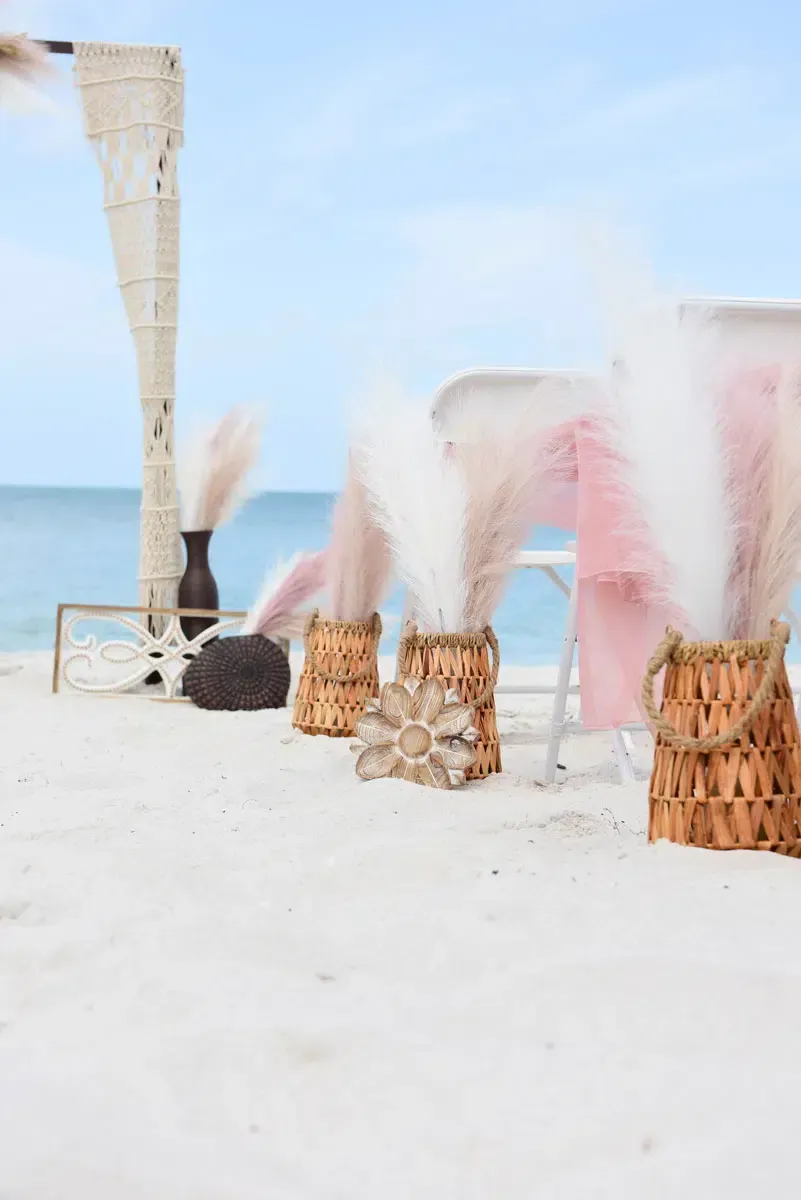 Beach wedding decorations: wicker baskets, pampas grass, driftwood on white sand, with ocean backdrop.