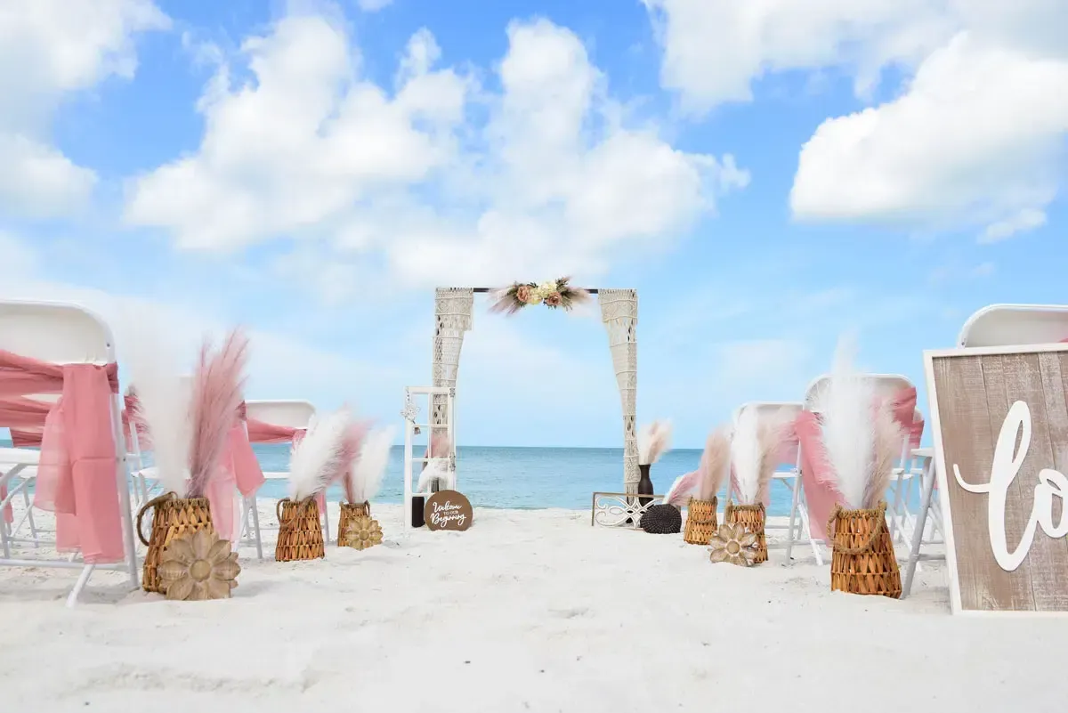 Beach wedding ceremony setup with arch, chairs, and decorations on a white sandy beach, under a blue sky.