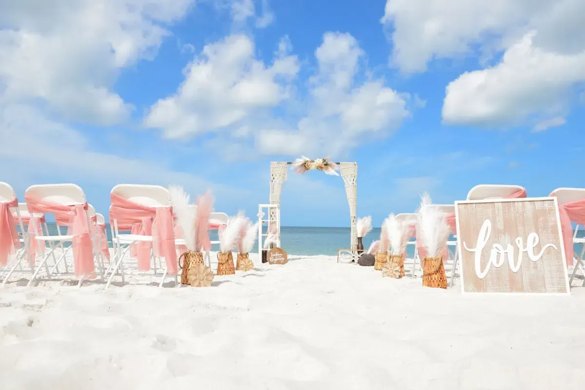 Beach wedding setup: white chairs with pink sashes, arch with flowers, 