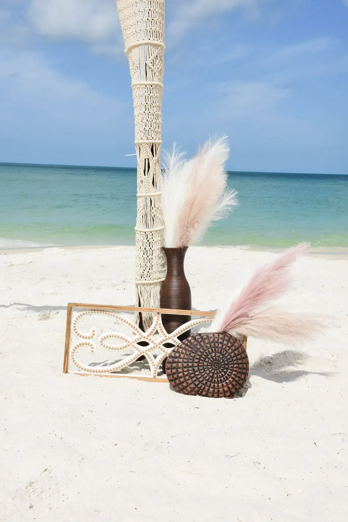 Beach scene with macrame column, vases of pink pampas grass, and decorative wooden elements.