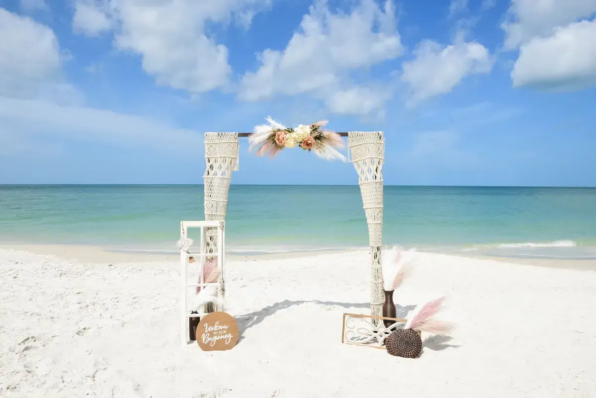Wedding arch on a white sand beach with ocean and blue sky in the background; decorated with flowers and pampas grass.