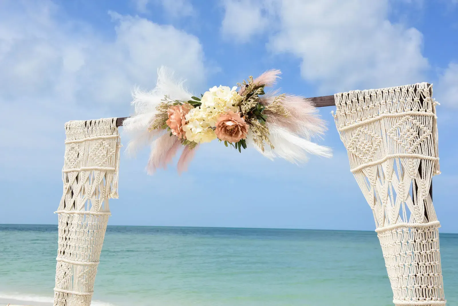 Beach wedding arch with macrame details, decorated with flowers and pampas grass, ocean background.