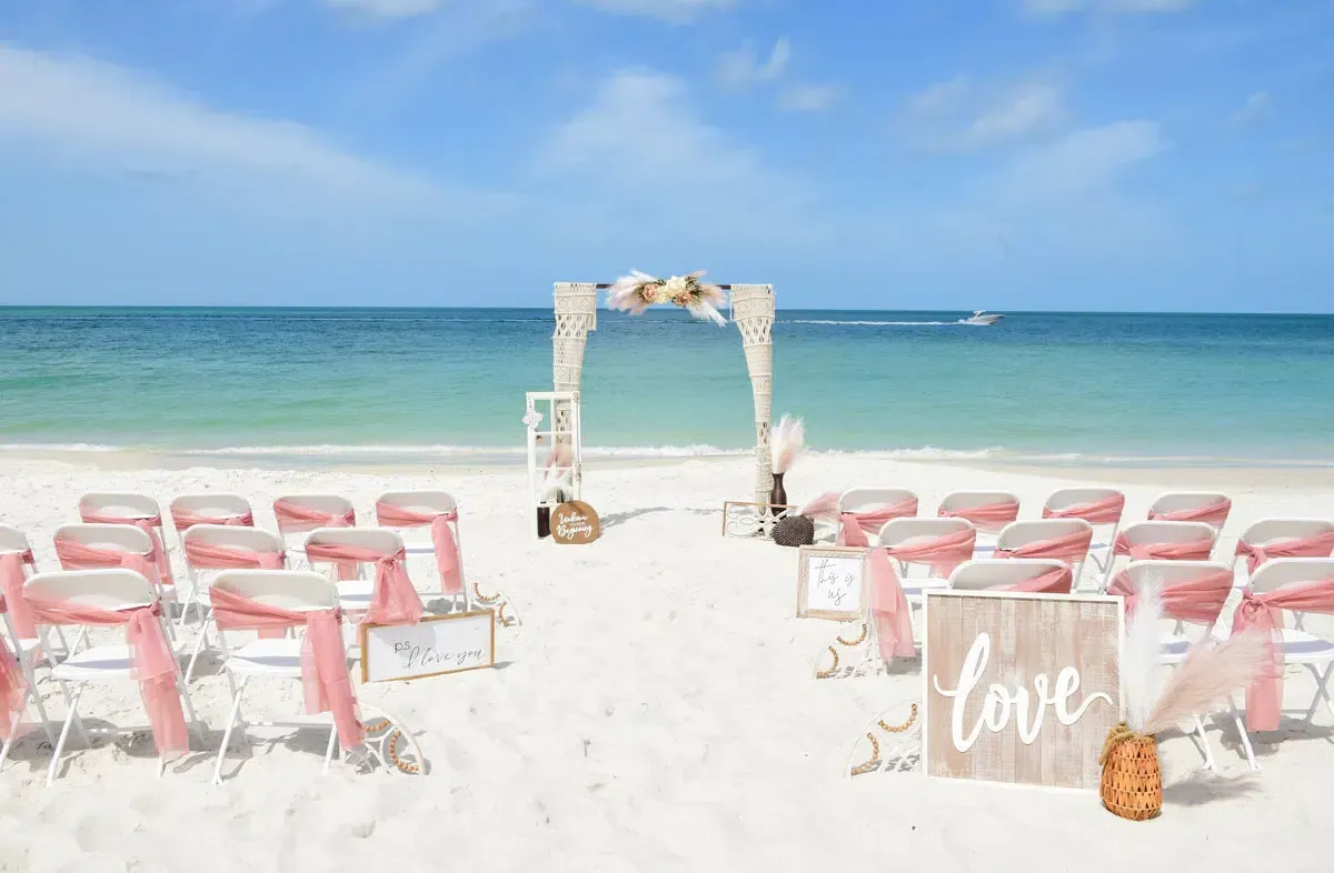 Beach wedding setup: white chairs with pink sashes face a decorated arch on white sand. Turquoise ocean and blue sky in background.