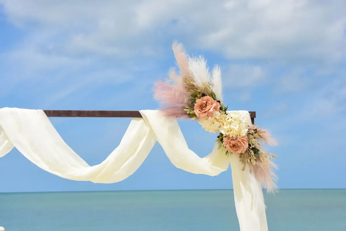 Wedding arch decorated with flowers and fabric, set on a beach with blue sky and ocean.