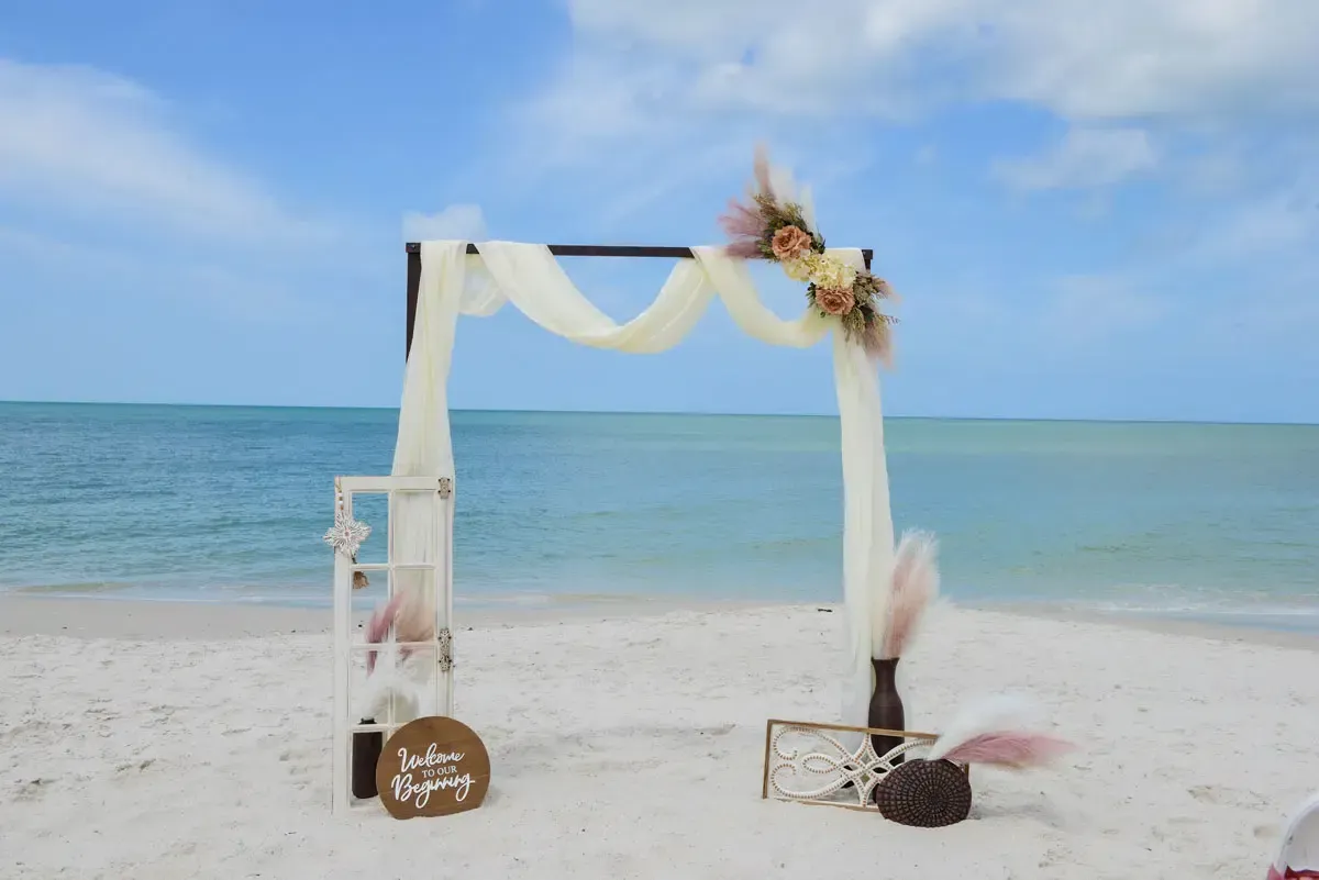 Wedding arch on a sandy beach decorated with fabric and flowers; the ocean in the background.