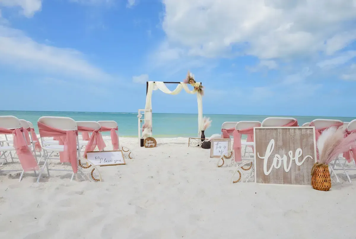 Beach wedding setup with white sand, blue water, and decorated chairs.