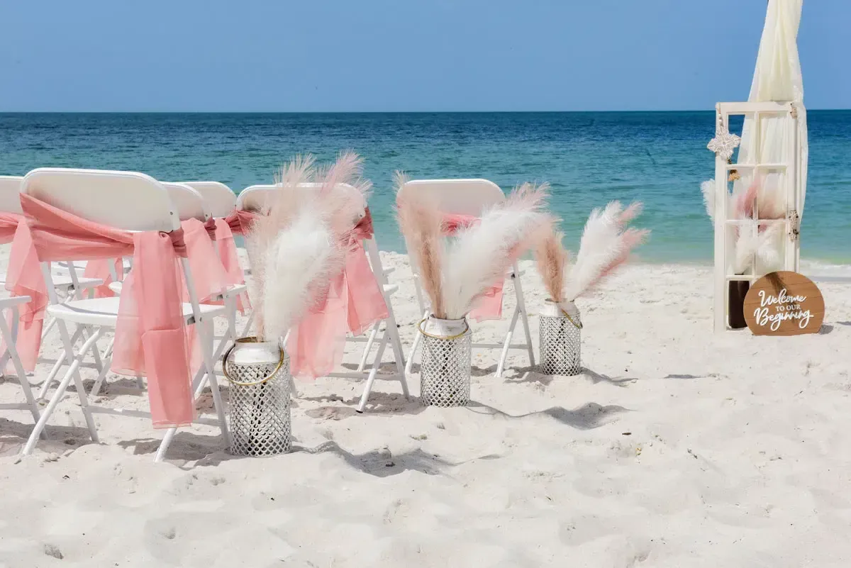Beach wedding ceremony setup: white chairs with pink sashes, pampas grass decor, ocean background, sunny day.