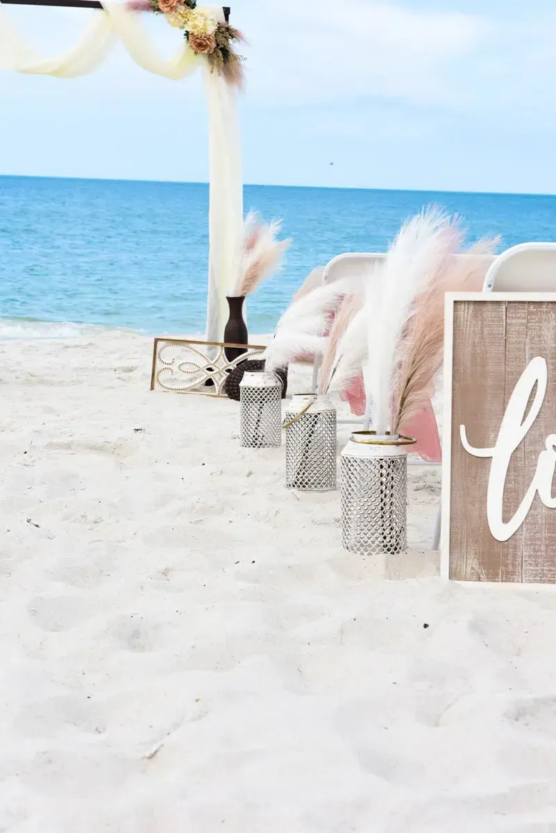 Beach wedding setup: white sand, ocean backdrop, arch decorated with flowers, lanterns, and a 