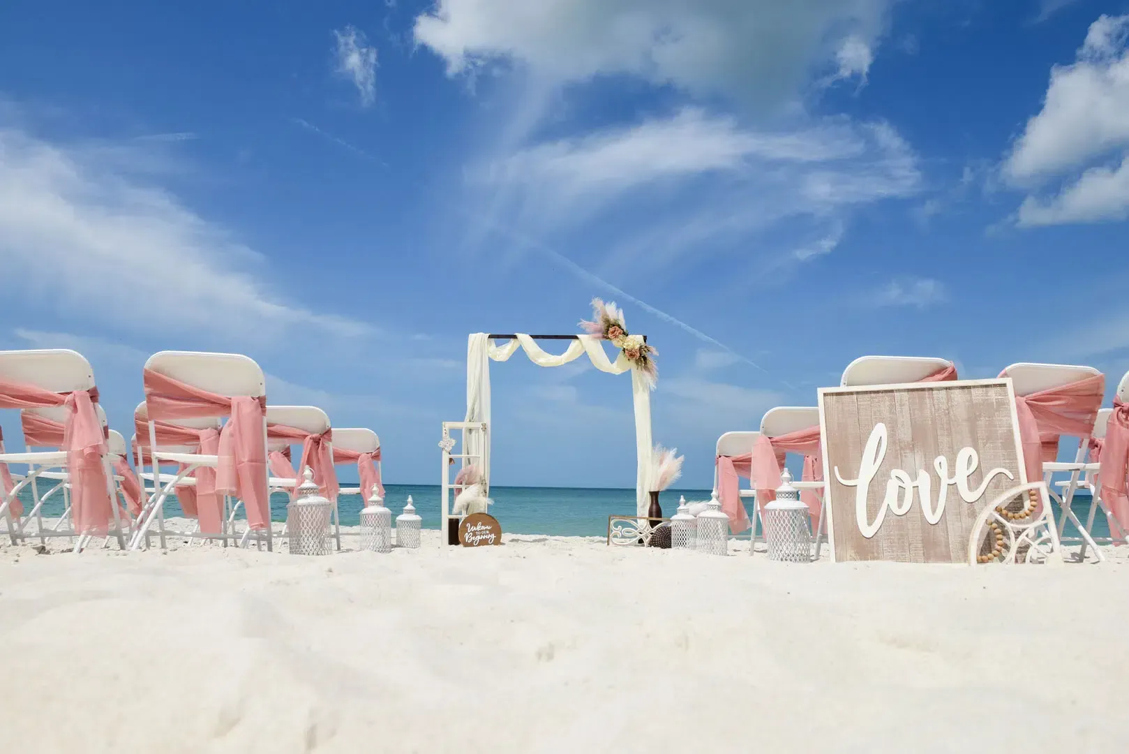 Beach wedding setup: white chairs with pink sashes, arch with draping and flowers, 