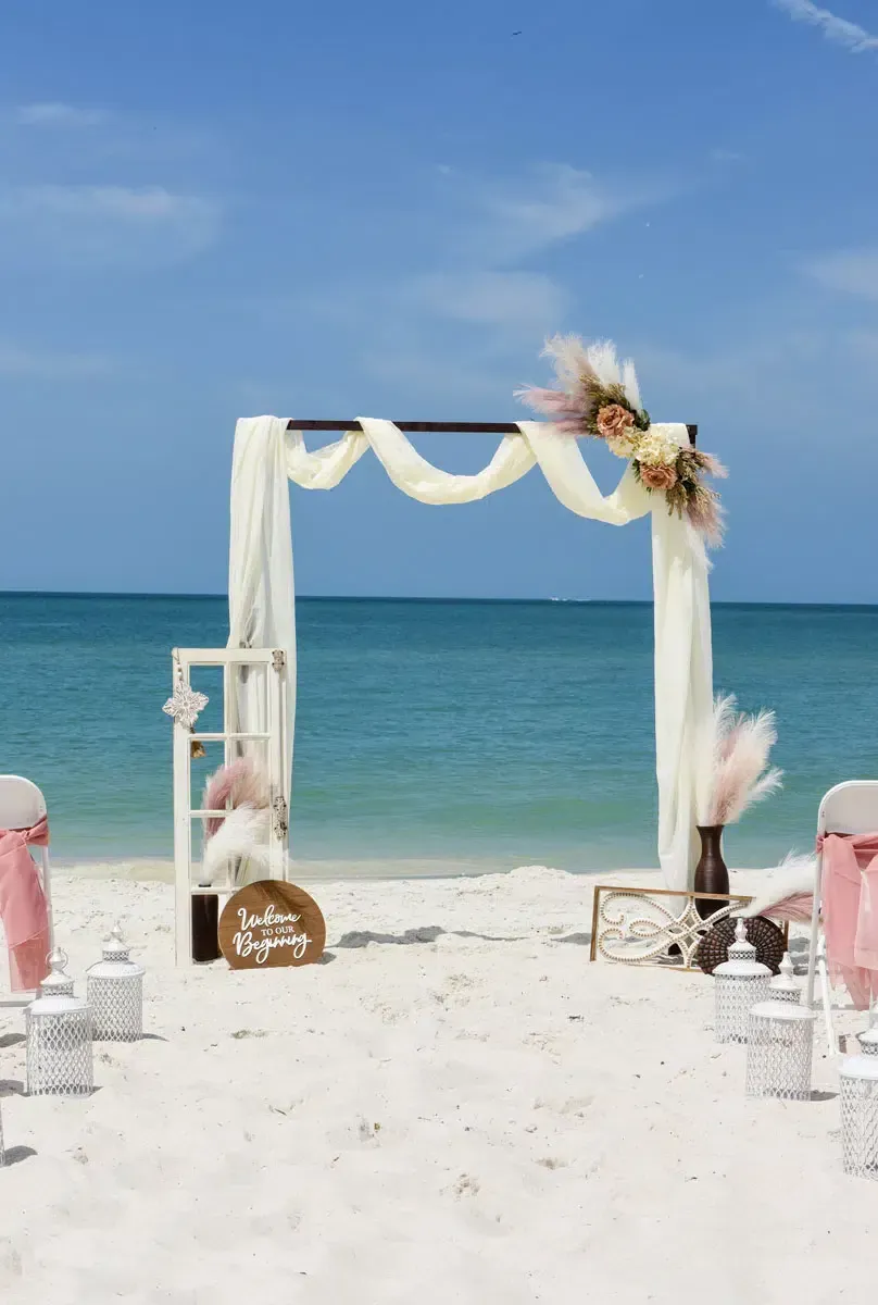 Beach wedding ceremony setup with white sand, blue water, and decorated arch.