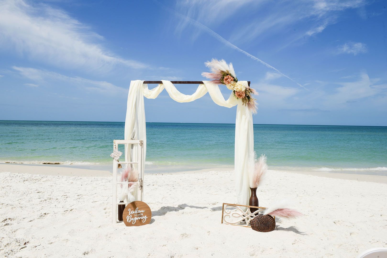Wedding arch on a white sand beach with ocean backdrop, decorated with flowers and sheer fabric.
