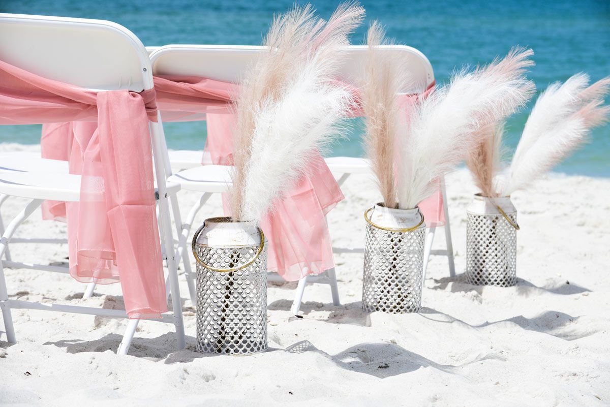 Wedding setup on beach with pink sashes on white chairs and pampas grass in silver vases.