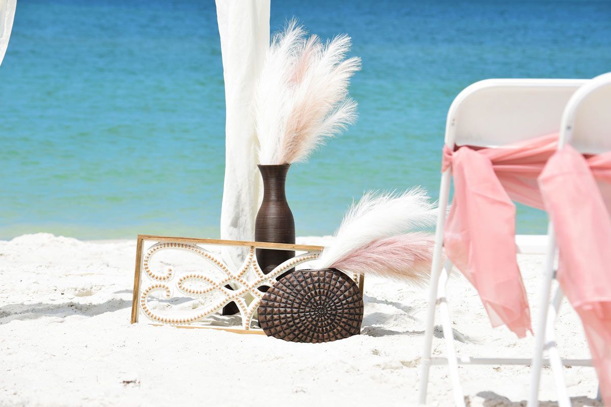 Beach wedding setup: white chairs with pink fabric, tall vases with feathery plumes, driftwood decor, and ocean backdrop.