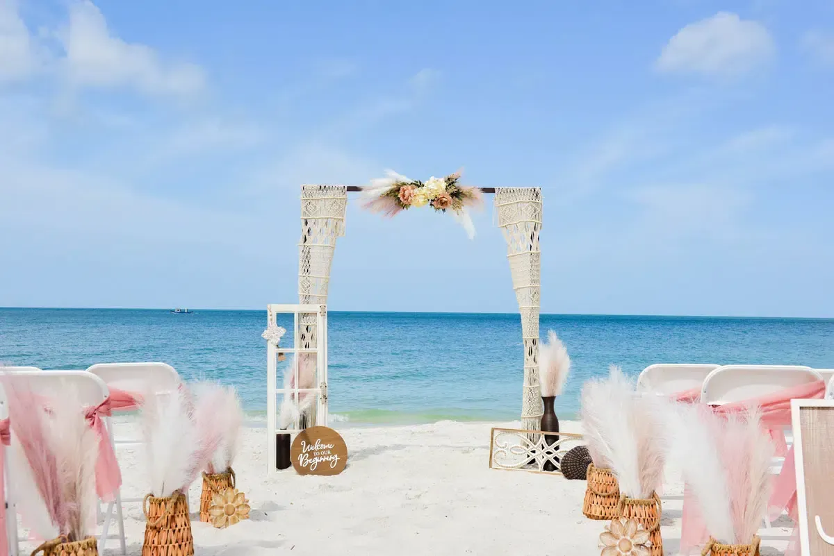 Beach wedding setup with arch, chairs, and decorations against a blue sky and ocean.