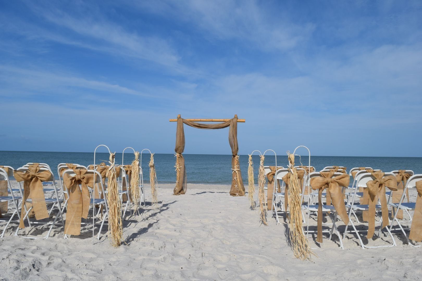 Beach wedding ceremony setup: white chairs with burlap accents face a bamboo arch against a blue sky.