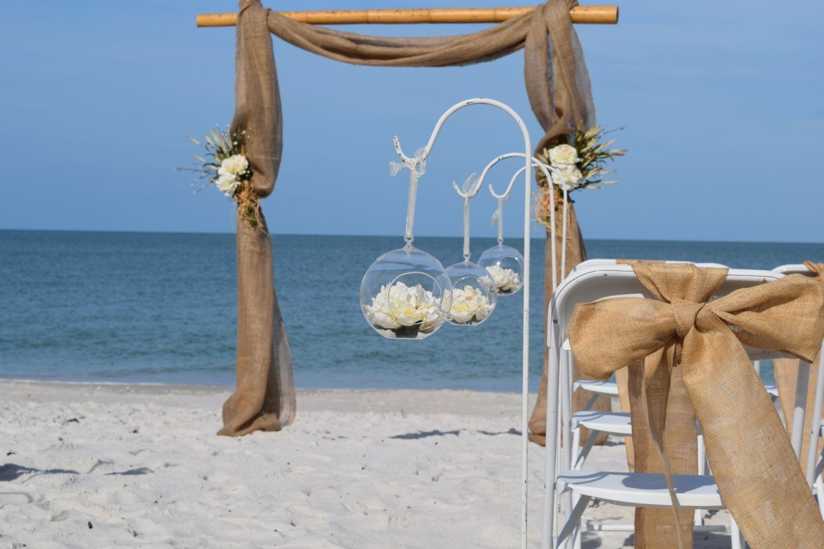 Beach wedding ceremony setup with white chairs, archway, and hanging floral globes.