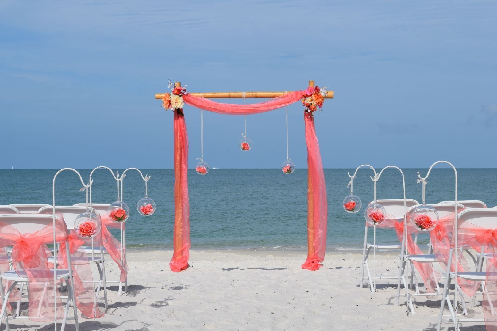 Beach wedding setup: arch with coral fabric and hanging flowers, white chairs with coral accents.
