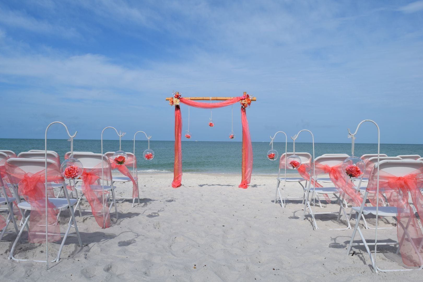 Beach wedding setup with chairs facing a decorated arch, ocean in background.