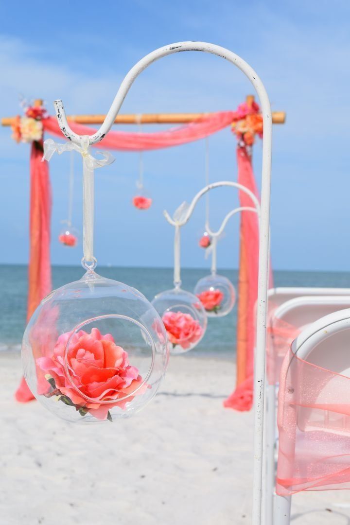 Beach wedding decor: Clear orbs with coral roses hang from a white stand, coral arch in background.
