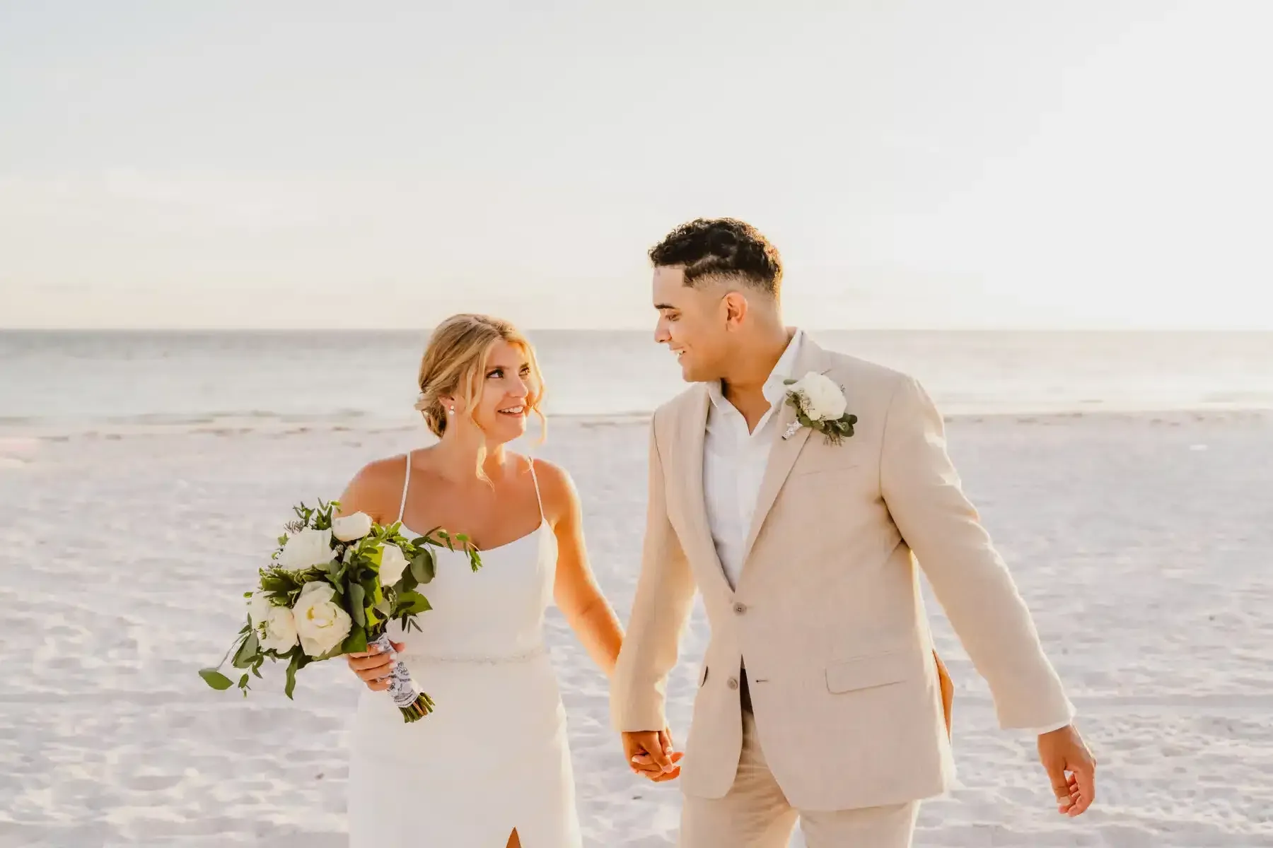 Newlyweds walking on a beach, holding hands. Bride smiles, holding bouquet. Groom in tan suit, looking at bride.