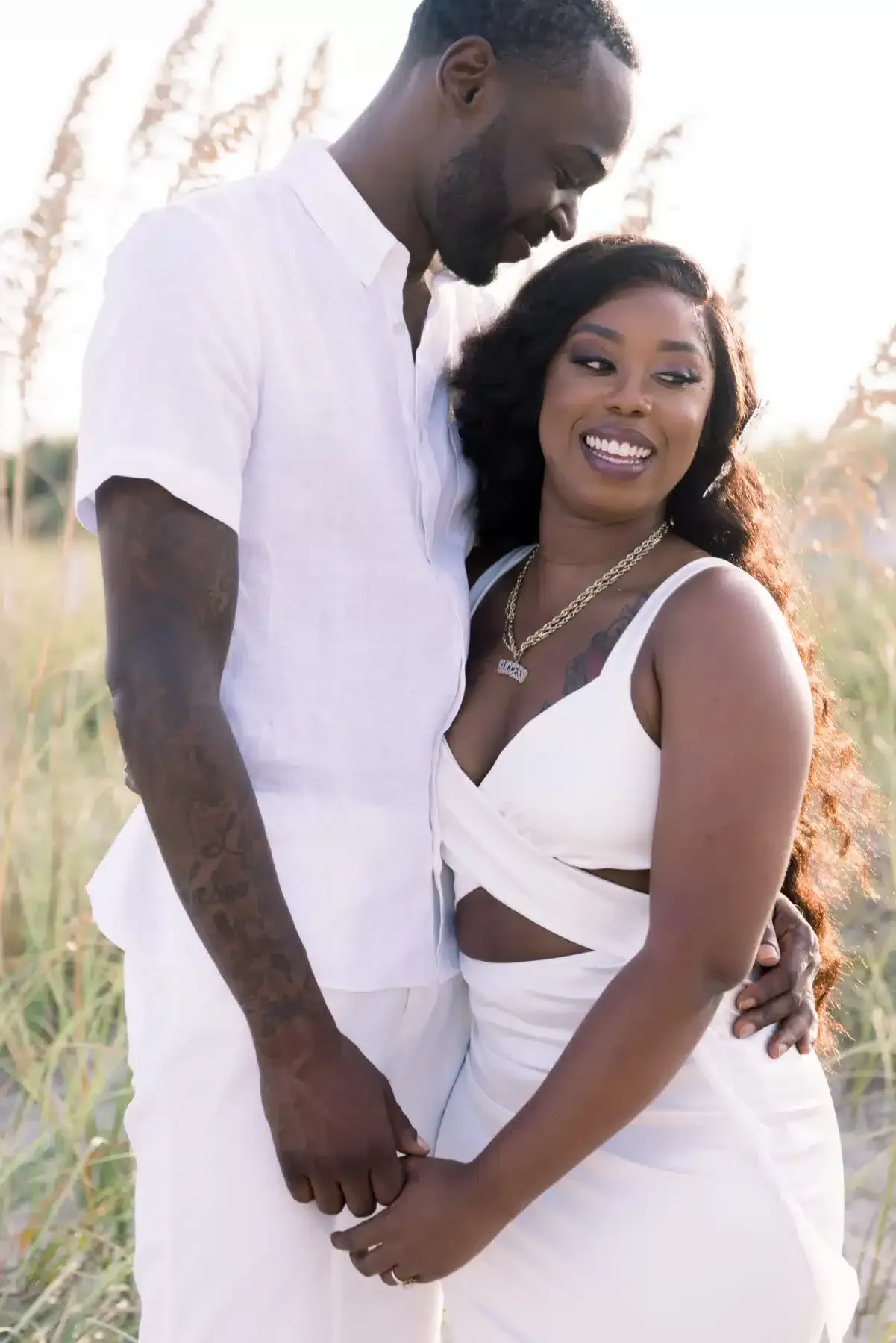 Couple embraces on a beach, both in white outfits. The woman smiles as they hold hands.